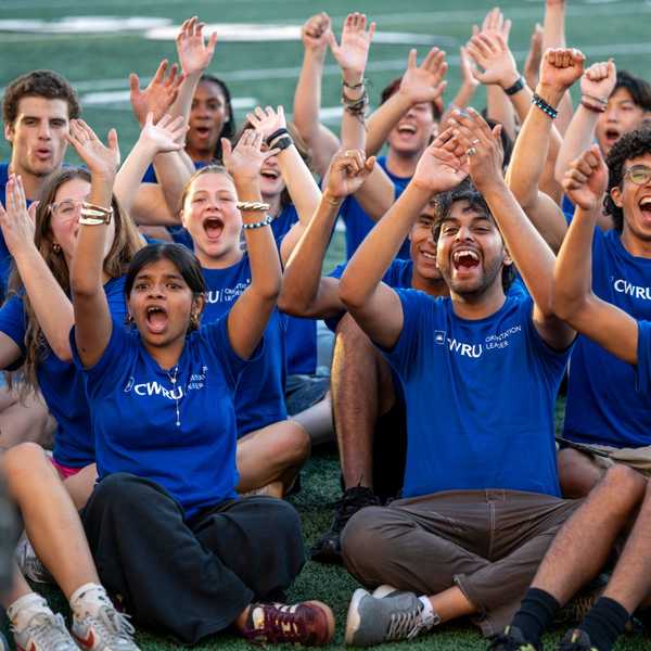 A group of CWRU orientation leaders sitting together on DiSanto Field in matching shirts, smiling and cheering.