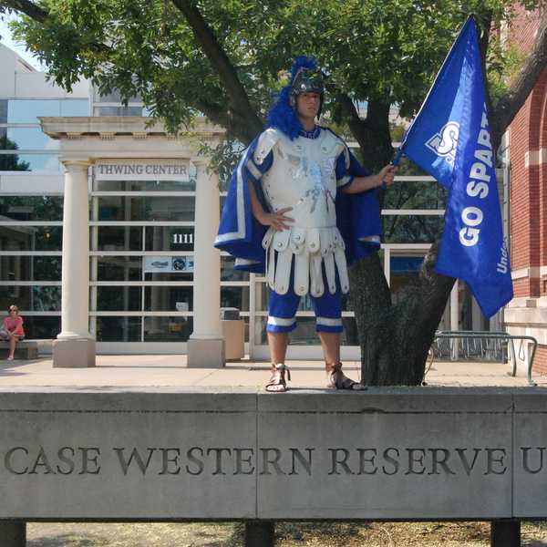 An older version of mascot Spartie posing with a blue flag that reads, “Go Spartans,” in front of Thwing Student Center.